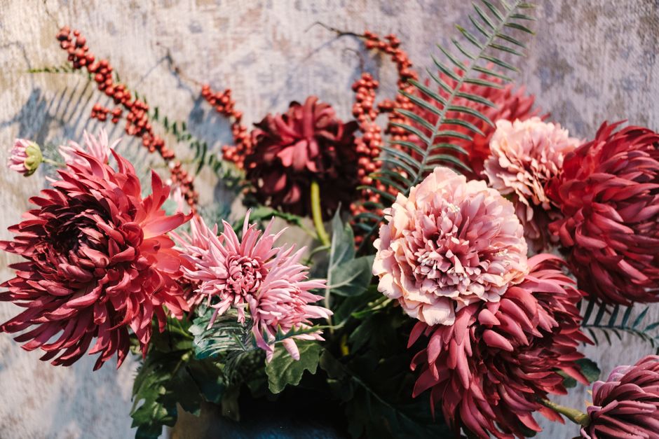 A vibrant floral arrangement featuring large, round, ruffled peonies in shades of soft blush pink and deep burgundy, complemented by spiky, pinkish-purple dahlias and sprigs of red berries. The bouque