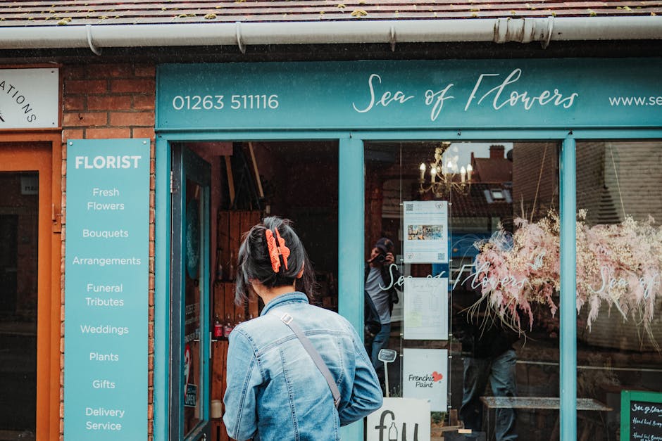 A woman wearing a denim jacket and a large red hair clip stands outside the flower shop 'Sea of Flowers', which features a bright teal sign with a list of services including fresh flowers, bouquets, a