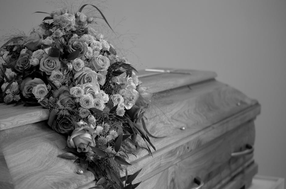 A large, elegant funeral floral arrangement resting on the edge of a wooden casket. The bouquet features a variety of flowers, including roses in shades of light and dark tones, complemented by smalle