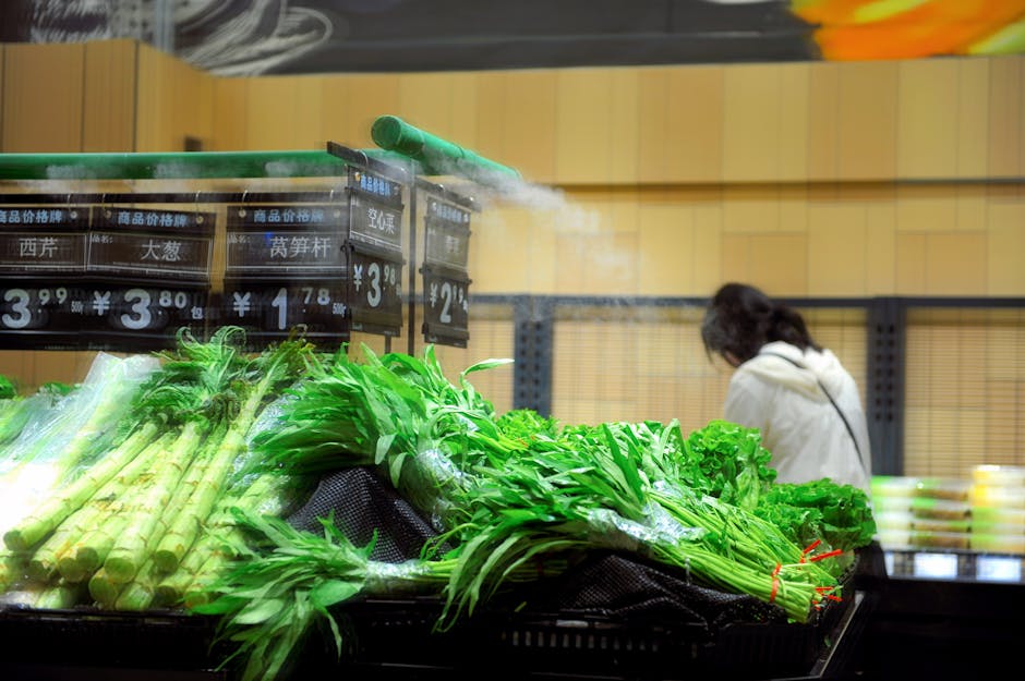 A display of fresh green vegetables including leeks and leafy greens arranged in black plastic baskets on a supermarket produce stand. In the background, a person in a white coat is shopping, with she