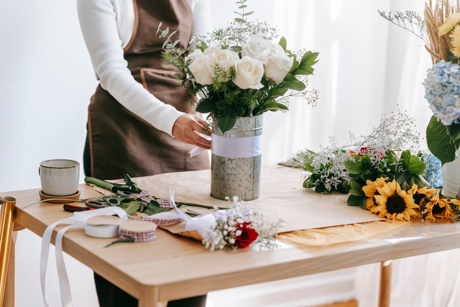A person in a brown apron and white long-sleeved top arranging a floral bouquet in a cylindrical grey marble-effect vase wrapped with a light purple ribbon, situated on a wooden table. The table holds