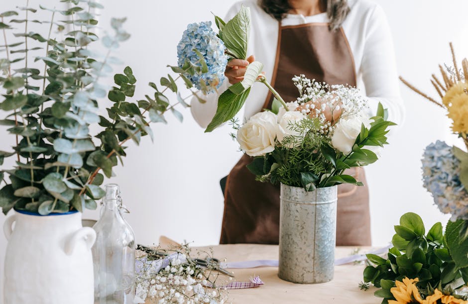 A person wearing a white shirt and brown apron arranging a bouquet of fresh flowers in a tall, silver cylindrical vase. The bouquet includes white roses, sprigs of baby's breath, and green foliage. Th