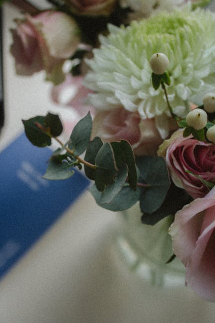 Close-up of a floral arrangement featuring soft pink roses, white chrysanthemums, and green eucalyptus leaves, arranged in a clear glass vase. The bouquet is positioned on a surface with natural light