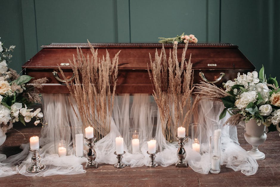 A floral arrangement featuring white roses, orchids, and cream-coloured blossoms with green foliage, positioned in white ceramic vases on each side of a polished wooden casket. Tall, dried pampas gras