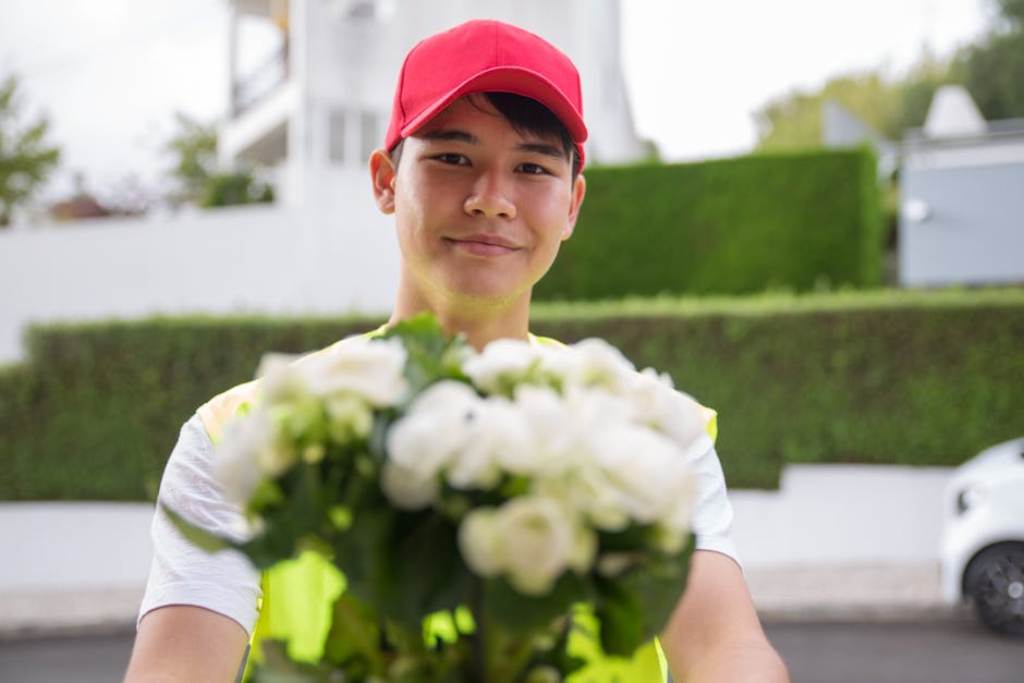 A delivery person wearing a red cap and yellow high-visibility vest is smiling and holding a bouquet of fresh, white flowers with green foliage, extending it towards the camera. The bouquet is arrange