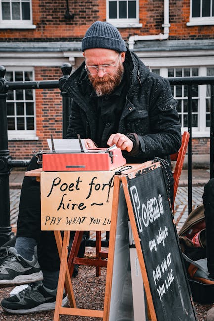 There is no image of flowers or floral arrangements in the provided photo. The image depicts a bearded man wearing a dark coat and knit cap, sitting outdoors at a wooden table on a city street. He is